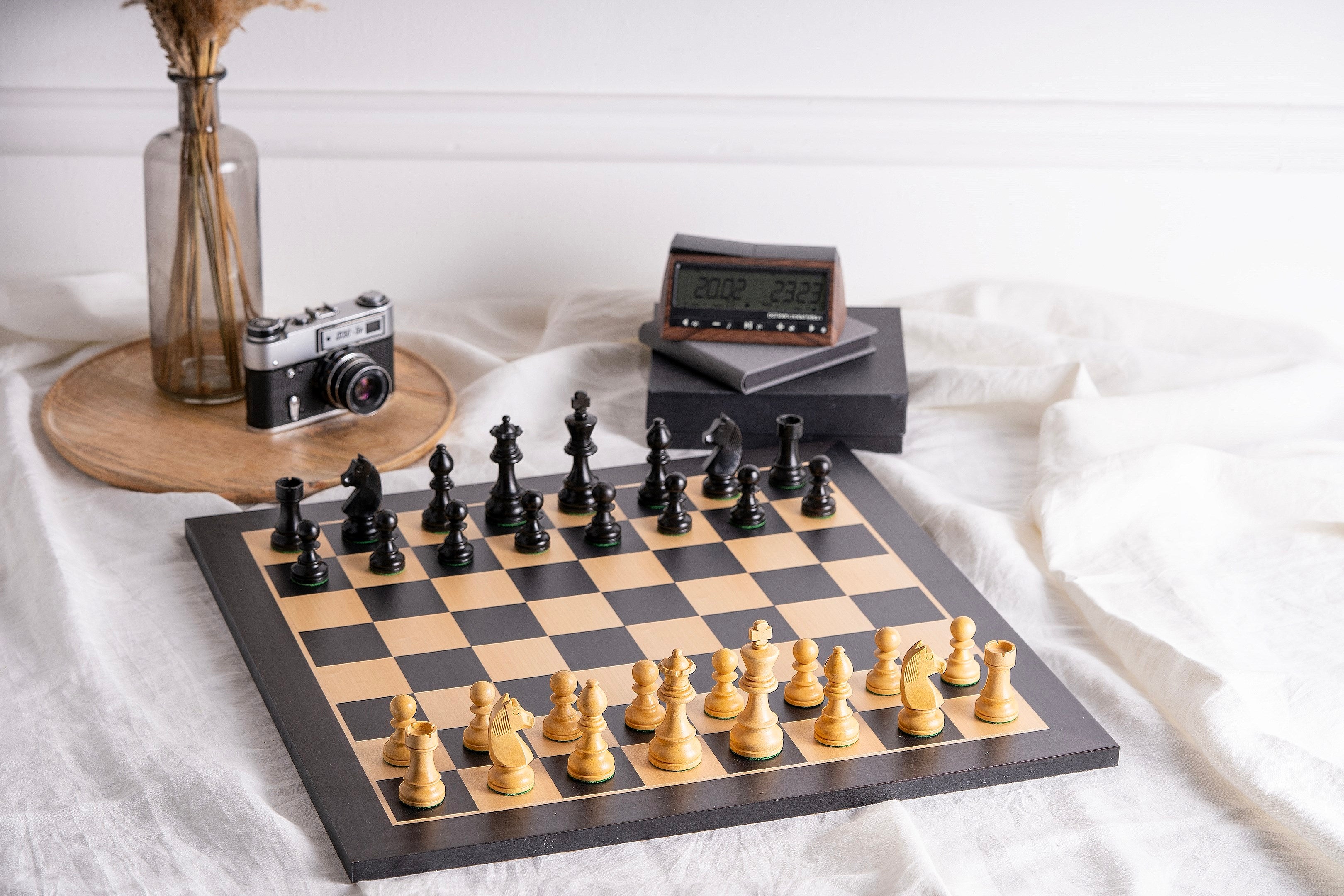 Échiquier & Pièces d'Échecs en Bois - Ensemble Staunton Noir et Blanc posé sur une nappe blanche, avec pièces d’échecs noires et blanches en bois, échiquier en bois noir et naturel, accompagné d’une pendule d’échecs et d’un appareil photo vintage en arrière-plan.