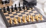 Charger l&#39;image dans la galerie, Zoom sur l’Échiquier &amp; Pièces d&#39;Échecs en Bois - Ensemble Staunton Noir et Blanc, avec pièces en bois sculpté noir et blanc sur un échiquier en bois clair et noir, ambiance élégante avec accessoires en arrière-plan.

