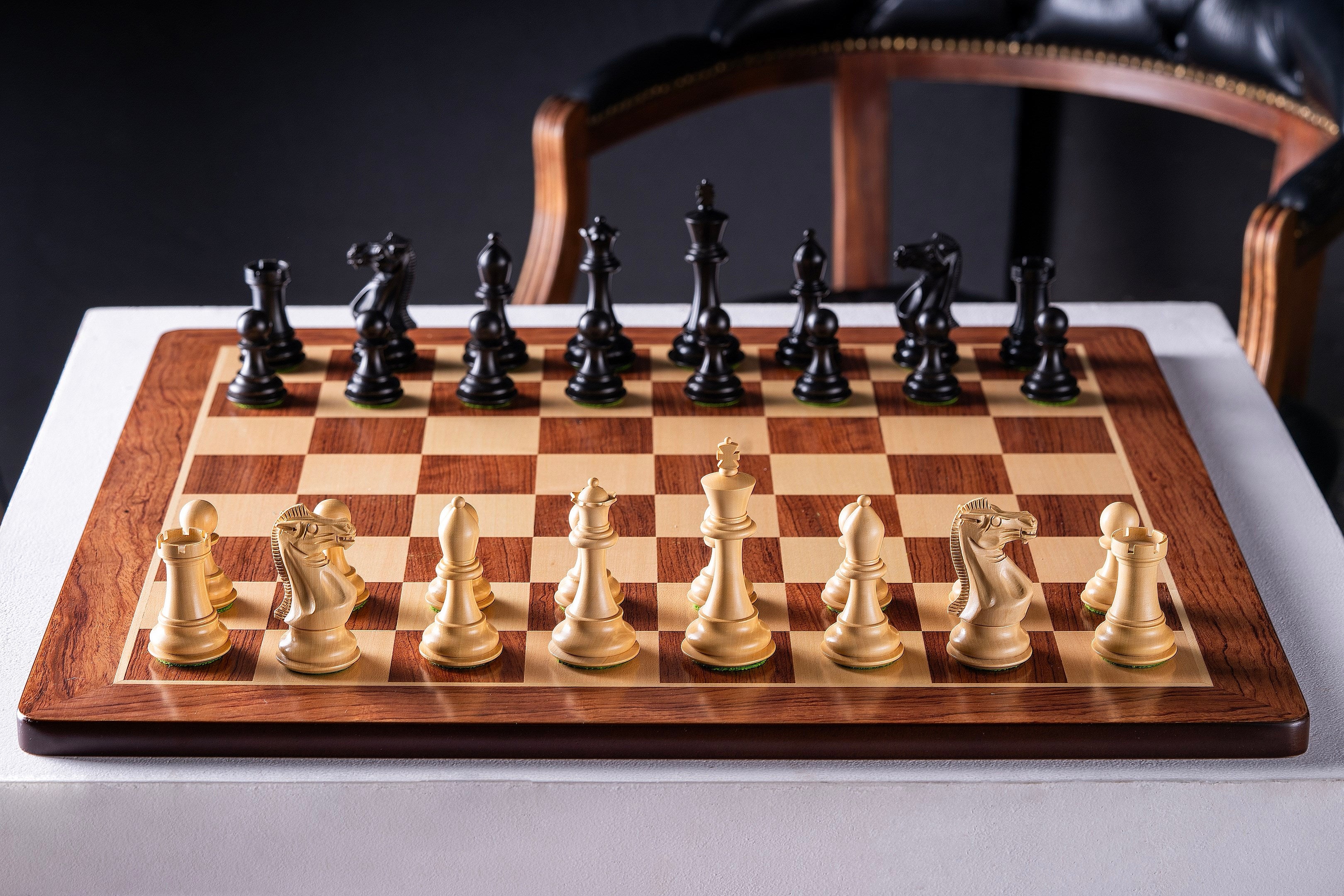 Échiquier en bois aux bords arrondis avec pièces d'échecs noires et blanches alignées, présenté sur une table blanche élégante.